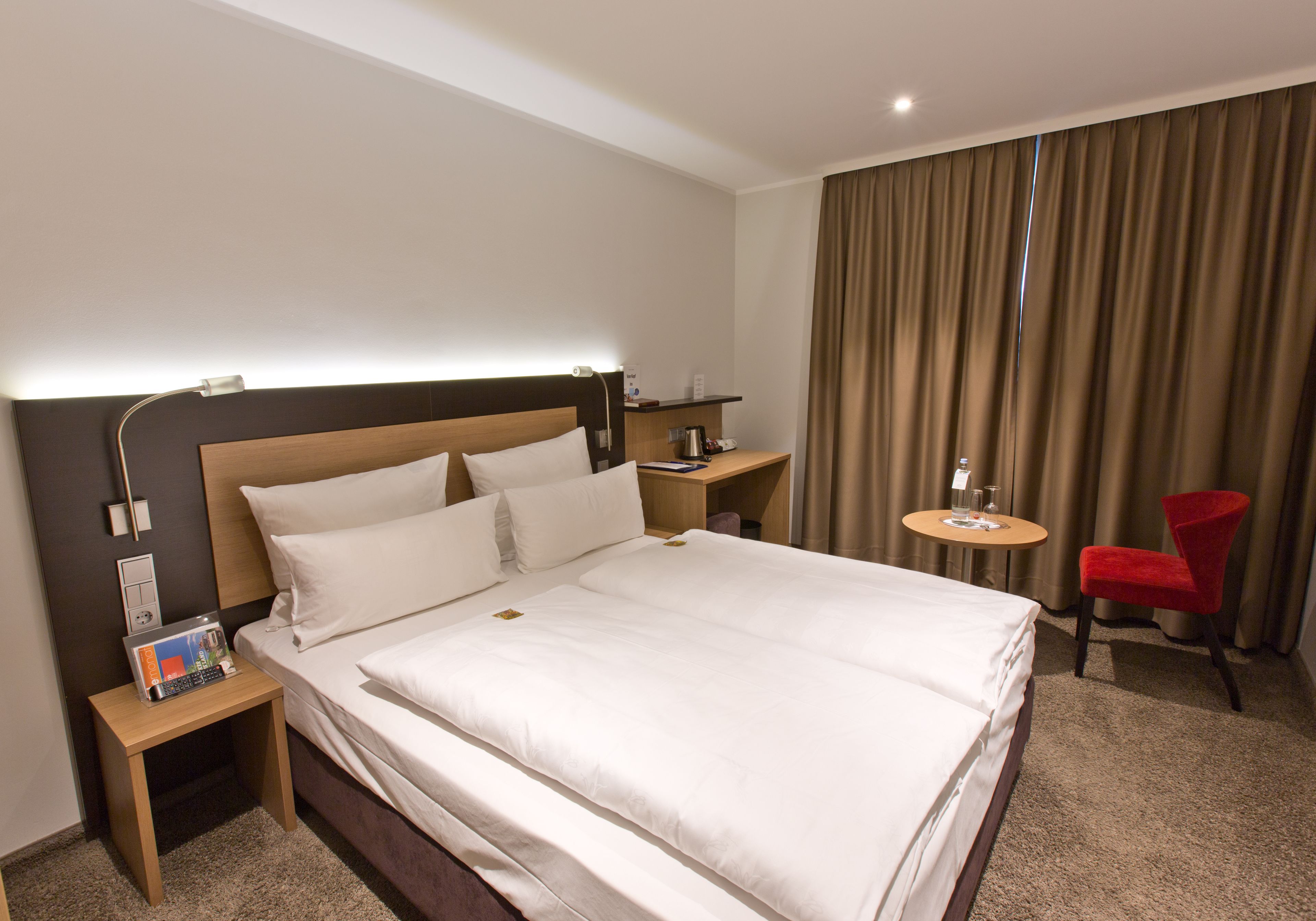 A hotel room with a large bed, a bedside table with a lamp, a small table with a chair, and a window with curtains.
