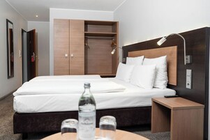 Una habitación de hotel moderna con una cama grande, un armario de madera, una mesita de noche con una lámpara y una botella de agua en la mesita.