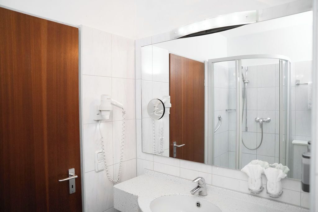 A modern bathroom with a white sink, a mirror, a shower, and a wooden door.