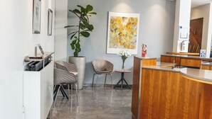Un espace d’accueil moderne avec un bureau en bois, un coin salon avec des fauteuils, une grande plante en pot et un paillasson.