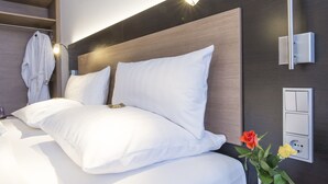 Une chambre d’hôtel moderne avec une tête de lit en bois, du linge de lit blanc, une table de chevet, un peignoir suspendu à un porte-manteau et un luminaire fixé au mur.