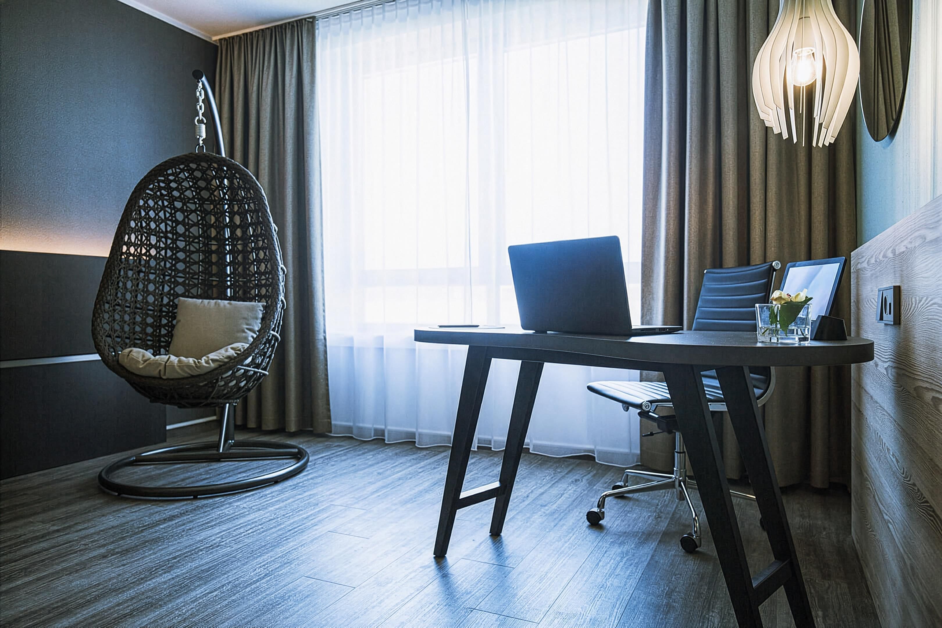 Une chambre moderne avec un ordinateur portable sur un bureau, un fauteuil suspendu et une grande fenêtre avec des rideaux.