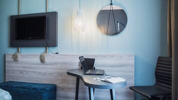 Una habitación moderna con una televisión de pantalla plana, una computadora portátil sobre una mesita pequeña, un espejo redondo y un otoman azul.