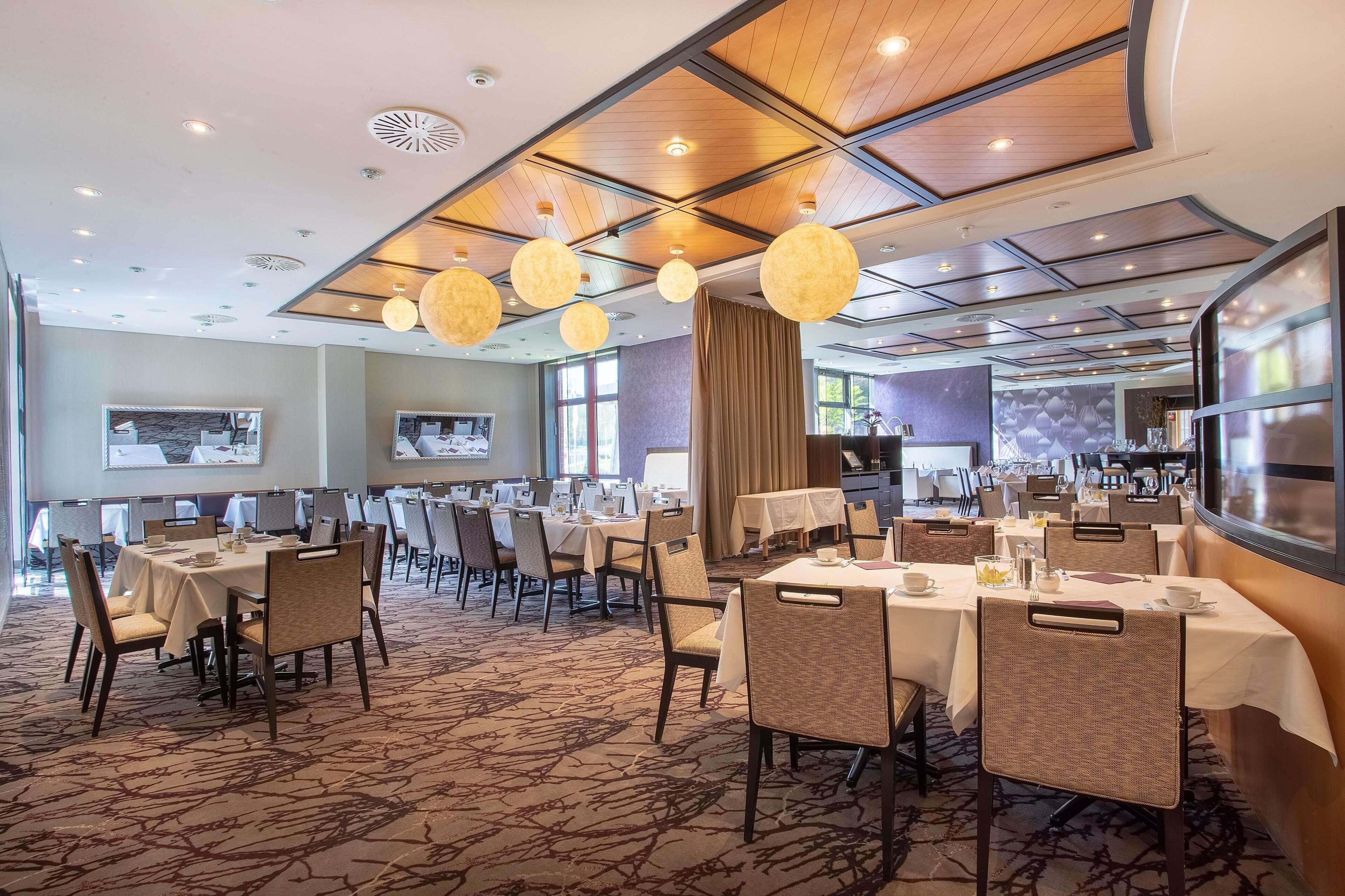 Ein Speisesaal eines Hotels mit mehreren Tischen und Stühlen, Deckenleuchten und einem gemusterten Teppich.