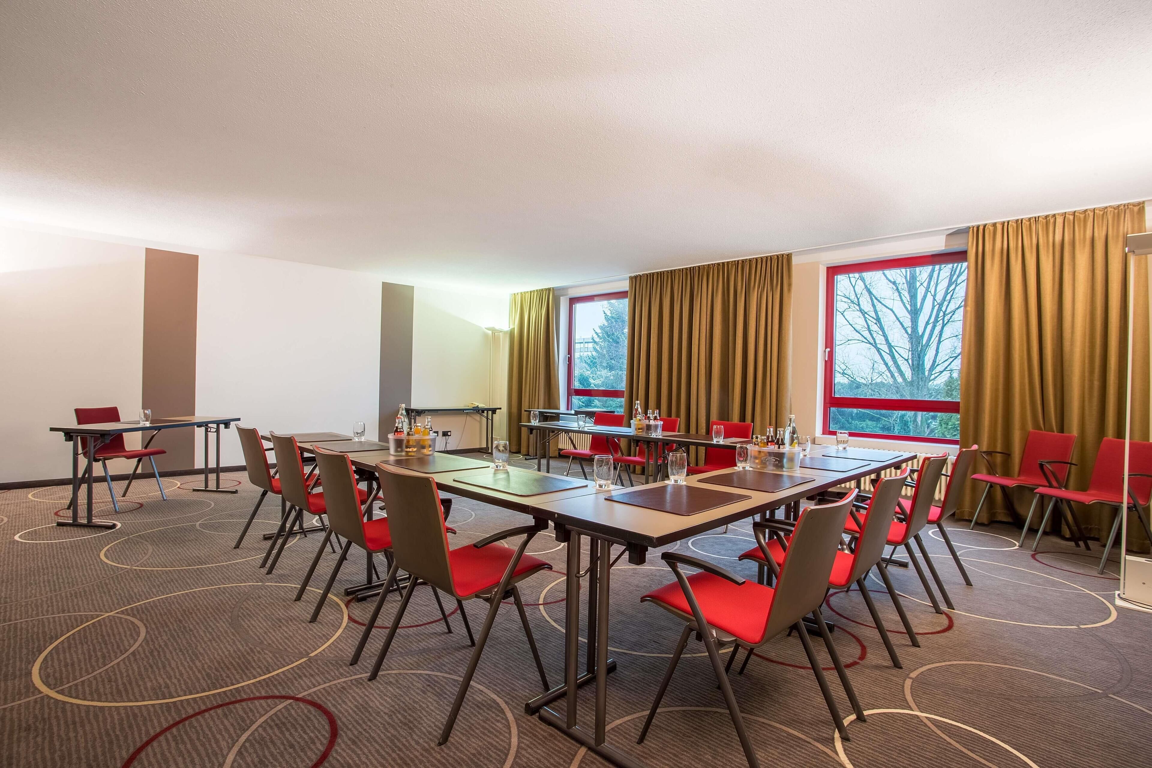 Ein Konferenzraum mit mehreren Tischen und Stühlen, großen Fenstern mit Vorhängen und einem Teppichboden.