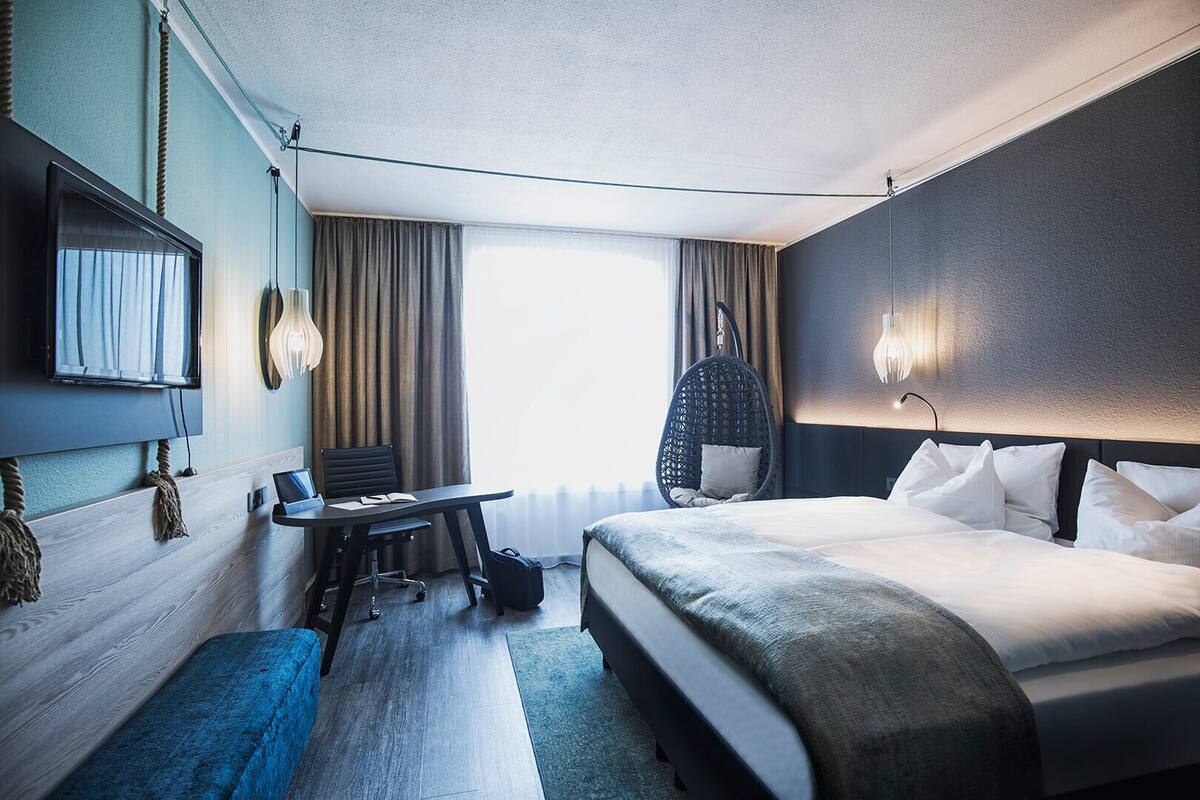 A hotel room with a bed, a desk, a chair, a hanging chair, and a TV.