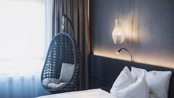 Ein Hotelzimmer mit Hängekorbschaukel, einem Bett mit weißer Bettwäsche, einer wandmontierten Lampe und Blick ins Freie durch ein Fenster mit Vorhängen.