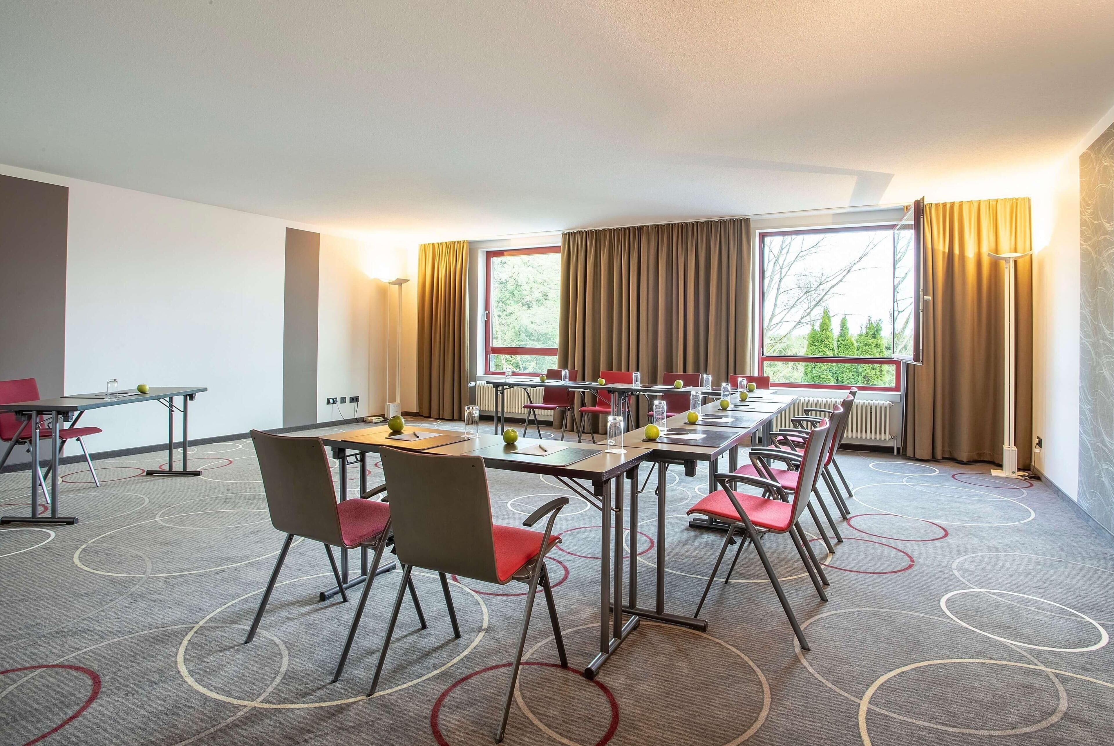 Un salón de conferencias con varias mesas y sillas, amplias ventanas con cortinas y piso alfombrado.