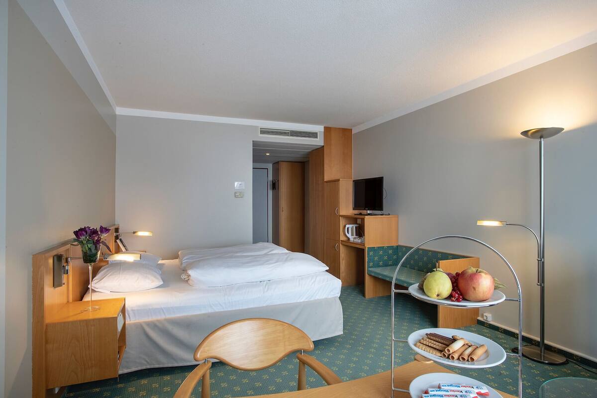 A hotel room with a bed, a desk, a chair, a TV, a lamp, and a tray table with food and drinks.