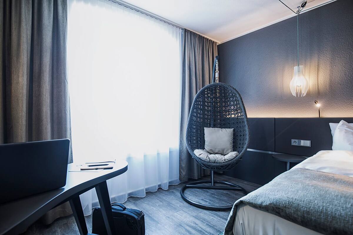 A hotel room with a bed, a desk with a laptop, a wicker chair, and a wall-mounted light fixture.