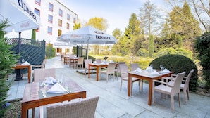 Een terras buiten met tafels, stoelen, parasols en een gebouw op de achtergrond.