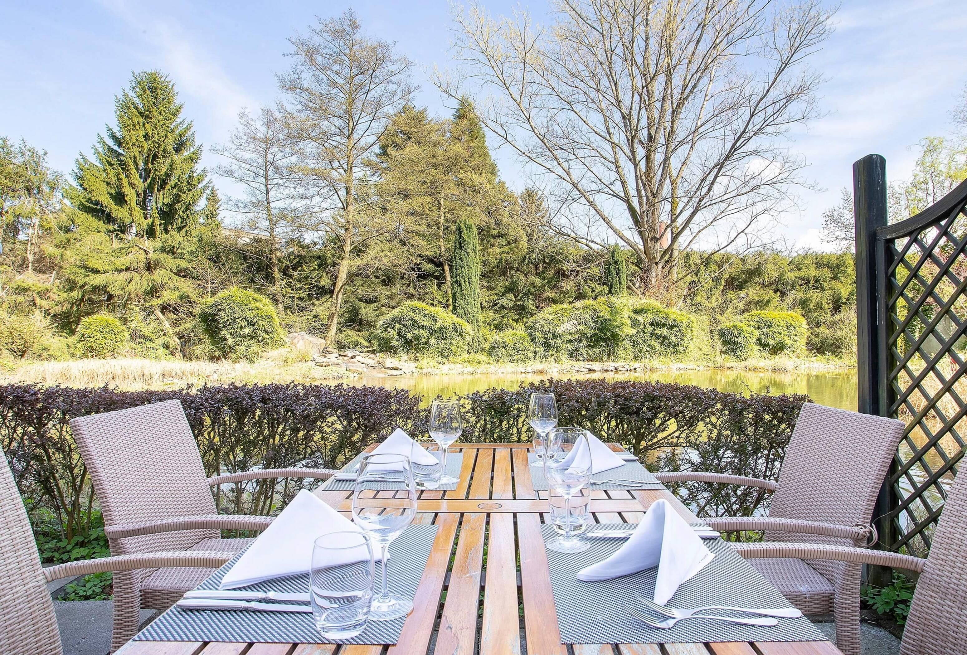 Mesa de comedor al aire libre con sillas de mimbre, dispuesta para dos personas, con vista a un estanque rodeado de árboles y arbustos.
