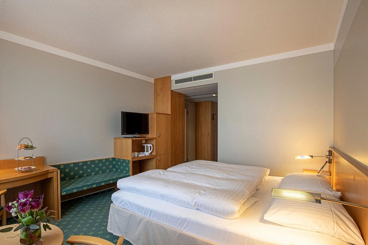 A hotel room with a bed, a TV, a small table with a tray, a bench, and a wall-mounted heater.