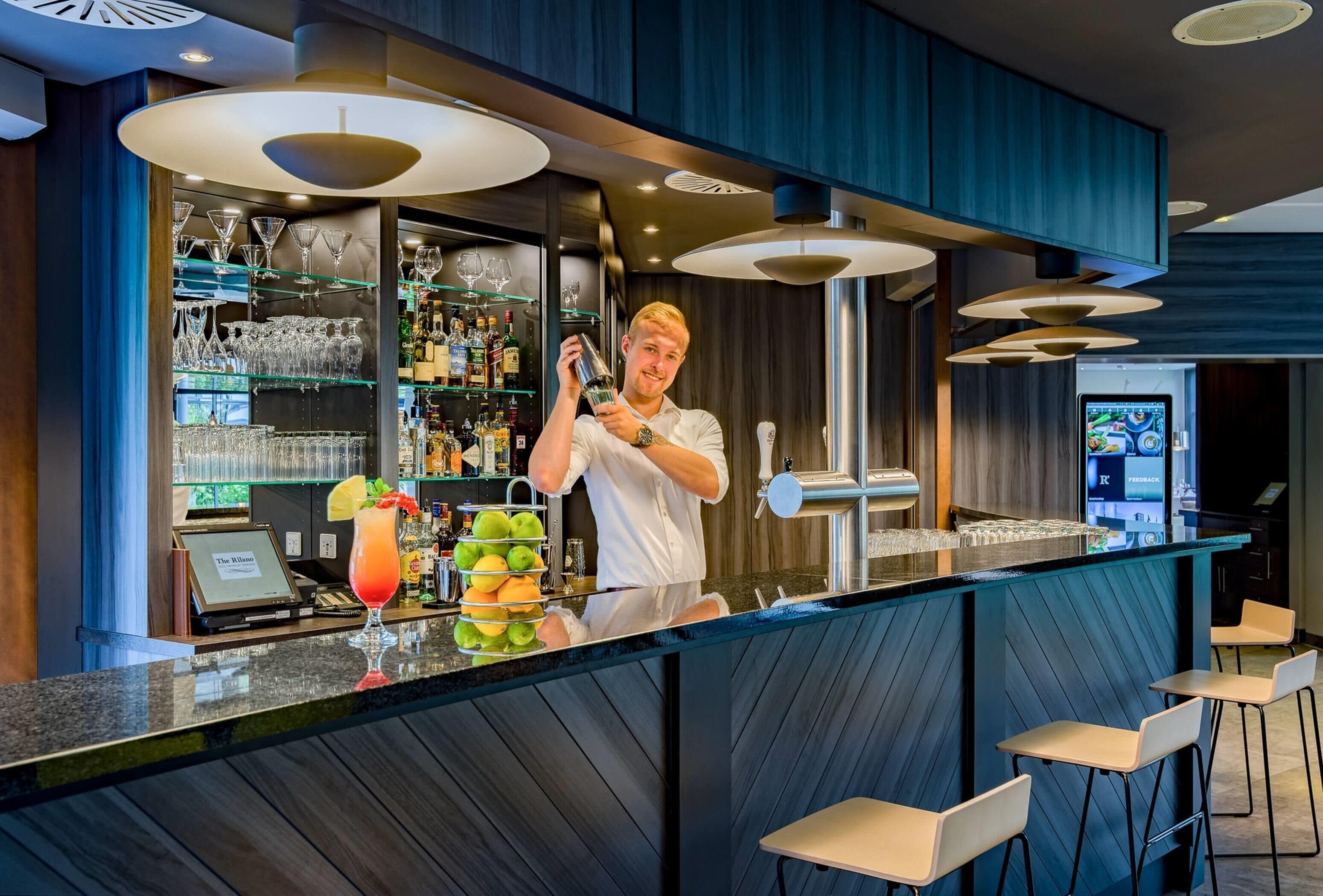 A bar area with a bartender, shelves of liquor bottles, a cocktail glass, and a digital menu screen.