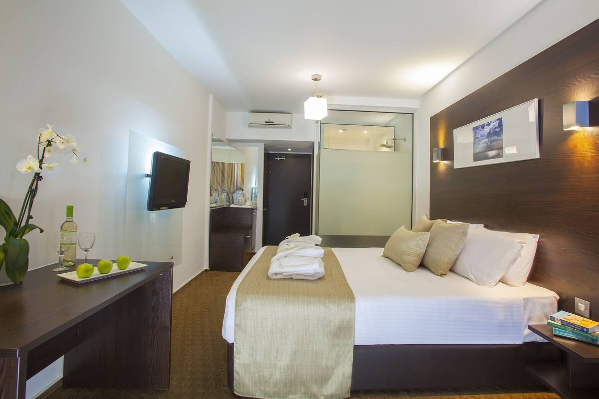 A hotel room with a bed, bedside table, TV, and a desk with a vase of flowers and a bottle of wine.