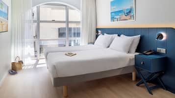 A hotel room with a large bed, a nightstand, a desk, and a view of the ocean through a window.