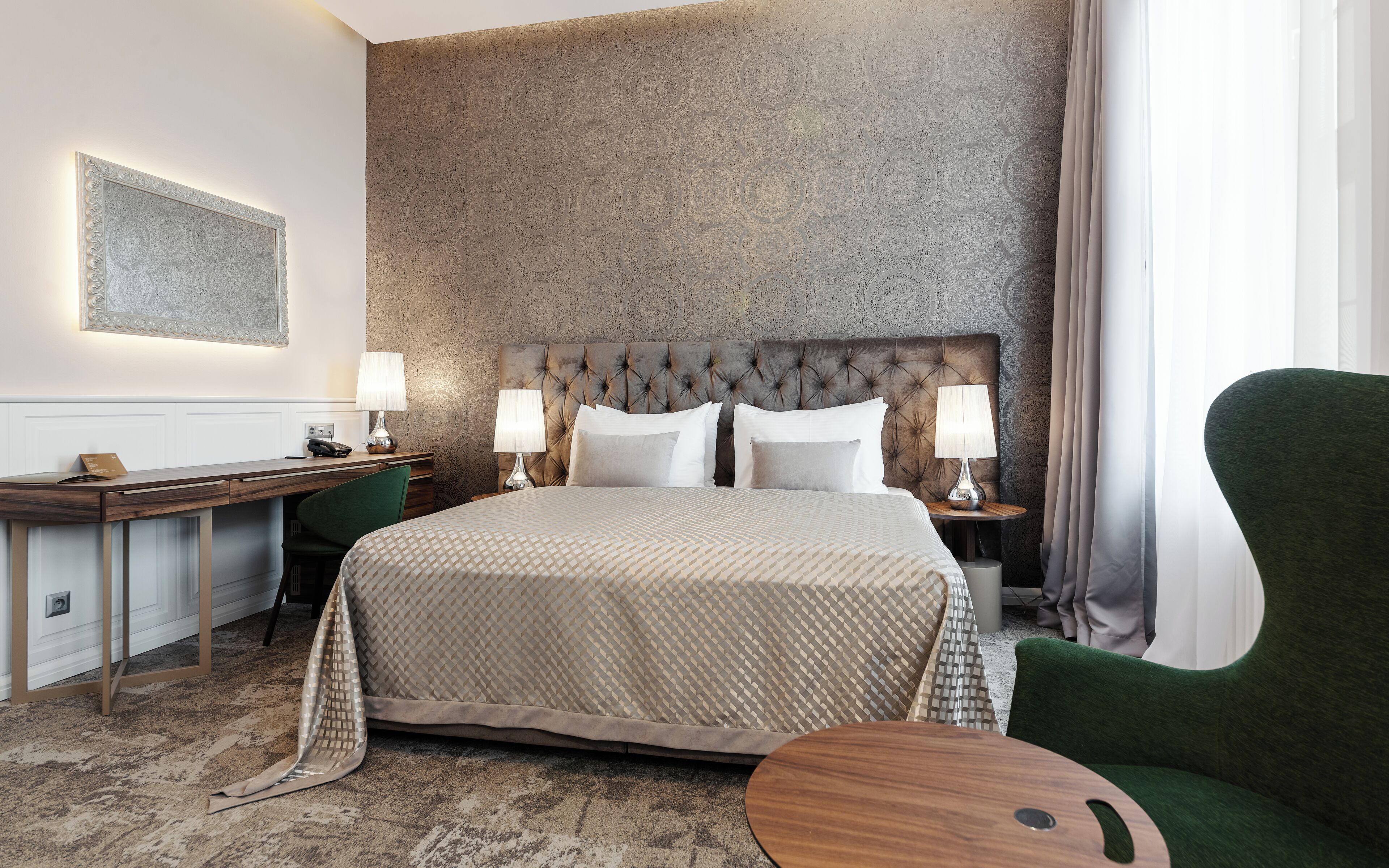 A hotel room with a large bed, two bedside tables with lamps, a desk with a telephone, and a green armchair.