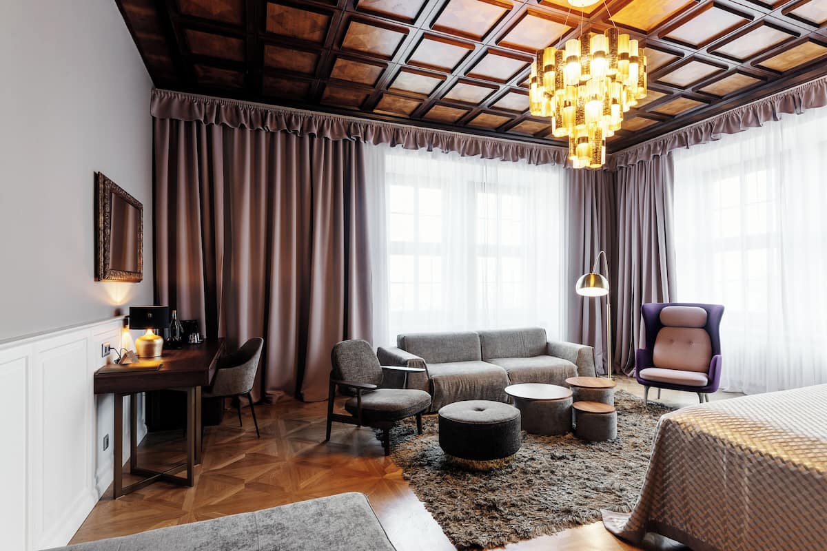 A modern hotel room with a wooden ceiling, a chandelier, a sofa, a chair, a desk, and a bed.