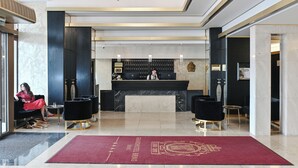 Un'area reception di un hotel con una scrivania, due sedie nere, un tappeto rosso e un lampadario.
