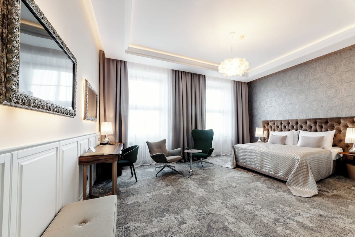 A hotel room with a large bed, a desk with a chair, a small table, and a green armchair.