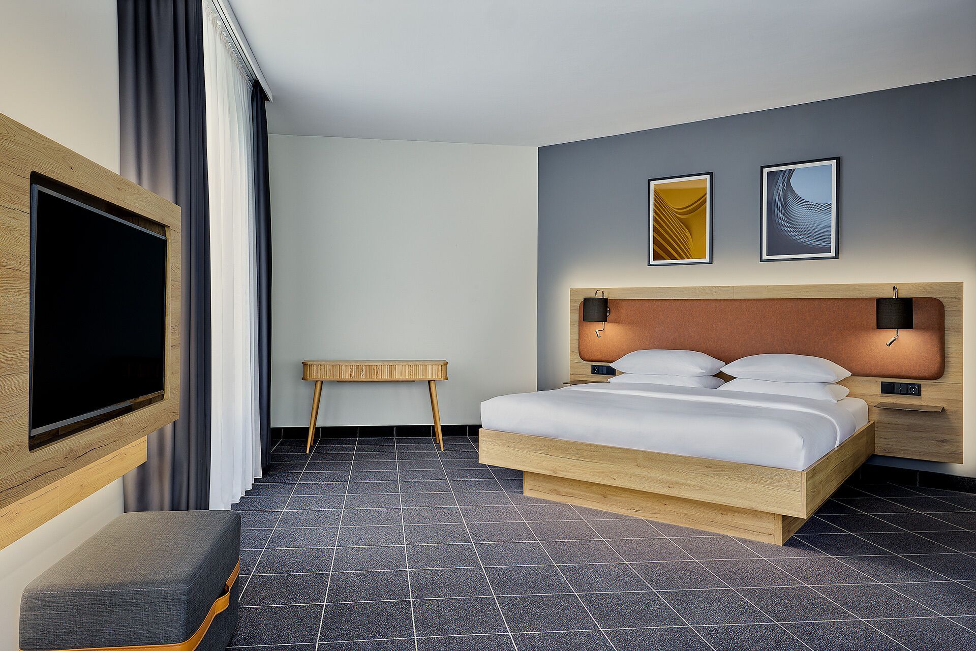 A modern hotel room with a large bed, a TV mounted on the wall, a small side table, and two framed artworks on the wall.