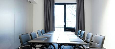 Un salón de conferencias con una larga mesa y sillas, ventanas amplias con cortinas y vistas al exterior.