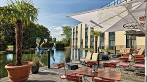 Un patio al aire libre con mesas y sillas, una sombrilla grande y vistas a un edificio a través de un canal.