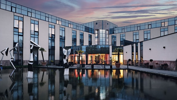 Un edificio moderno con piscina reflectante al frente, ventanas amplias y cielo despejado.