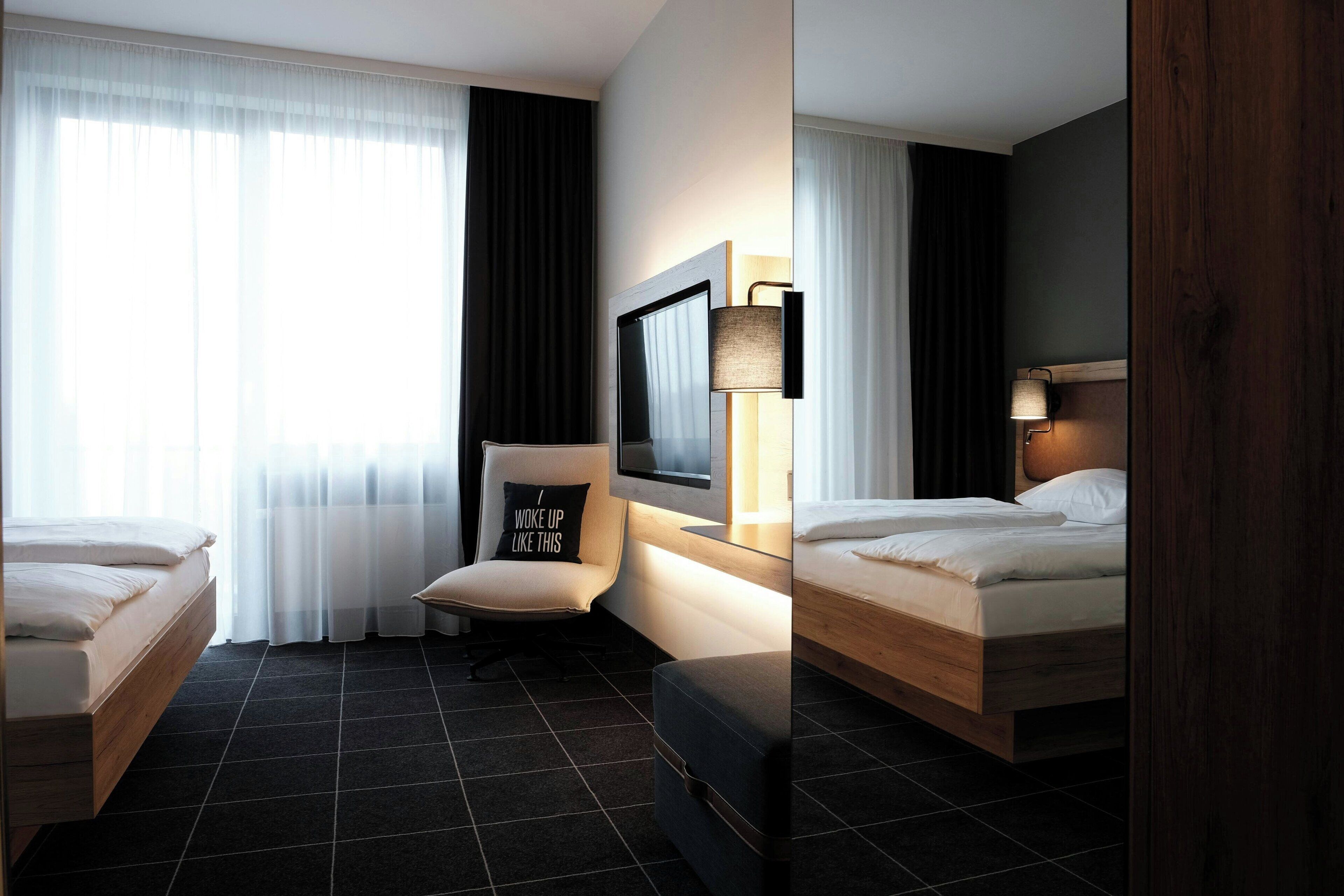 A hotel room with two beds, a chair with a pillow, a TV, and a lamp.