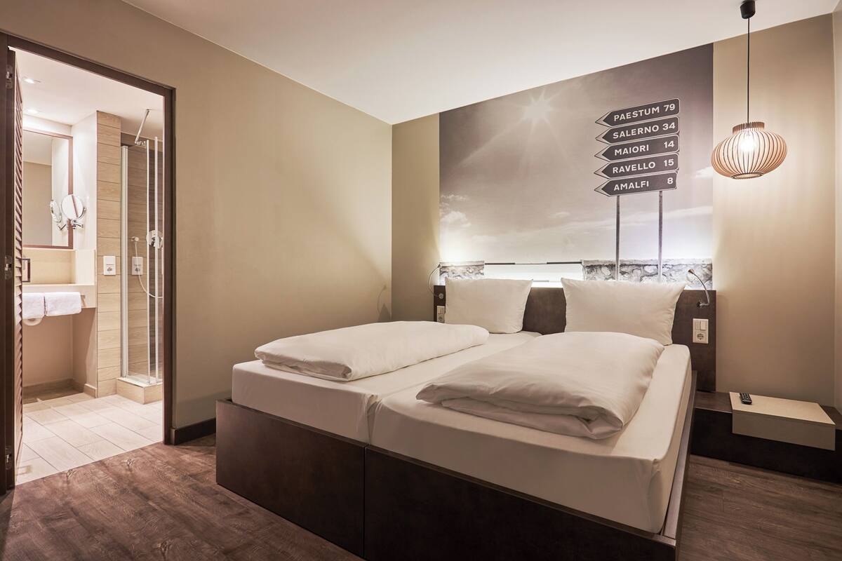 Una habitación de hotel con cama, mesita de noche, un cartel en la pared con indicaciones y una lámpara colgante.