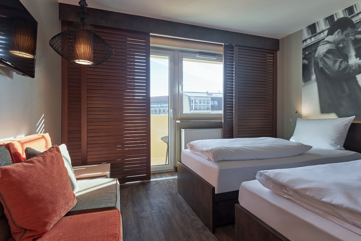 A hotel room with two beds, a seating area with a couch and pillows, a large window with blinds, and a wall-mounted artwork.