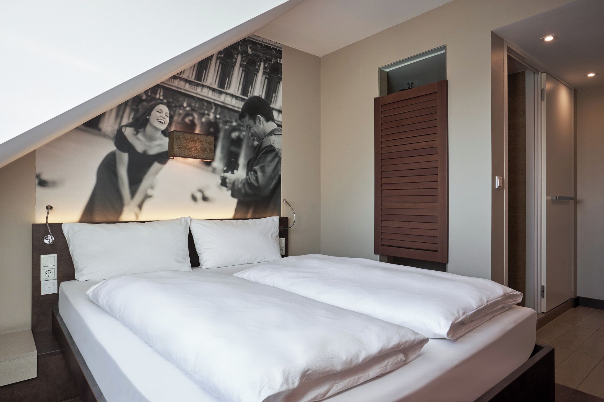 Une chambre d’hôtel moderne avec un grand lit, un téléviseur fixé au mur, une armoire en bois et une petite fenêtre.
