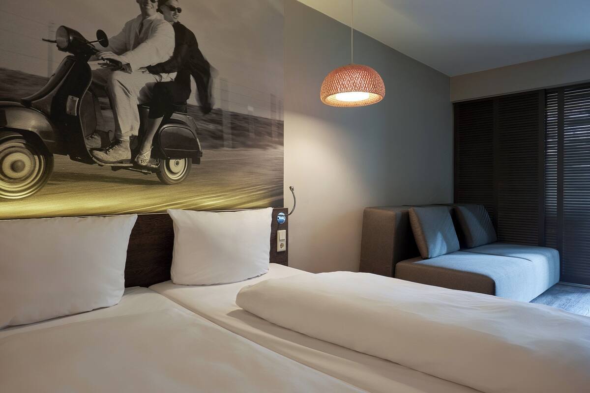 Habitación de hotel con cama, sofá, lámpara de pared y un mural de una scooter antigua.