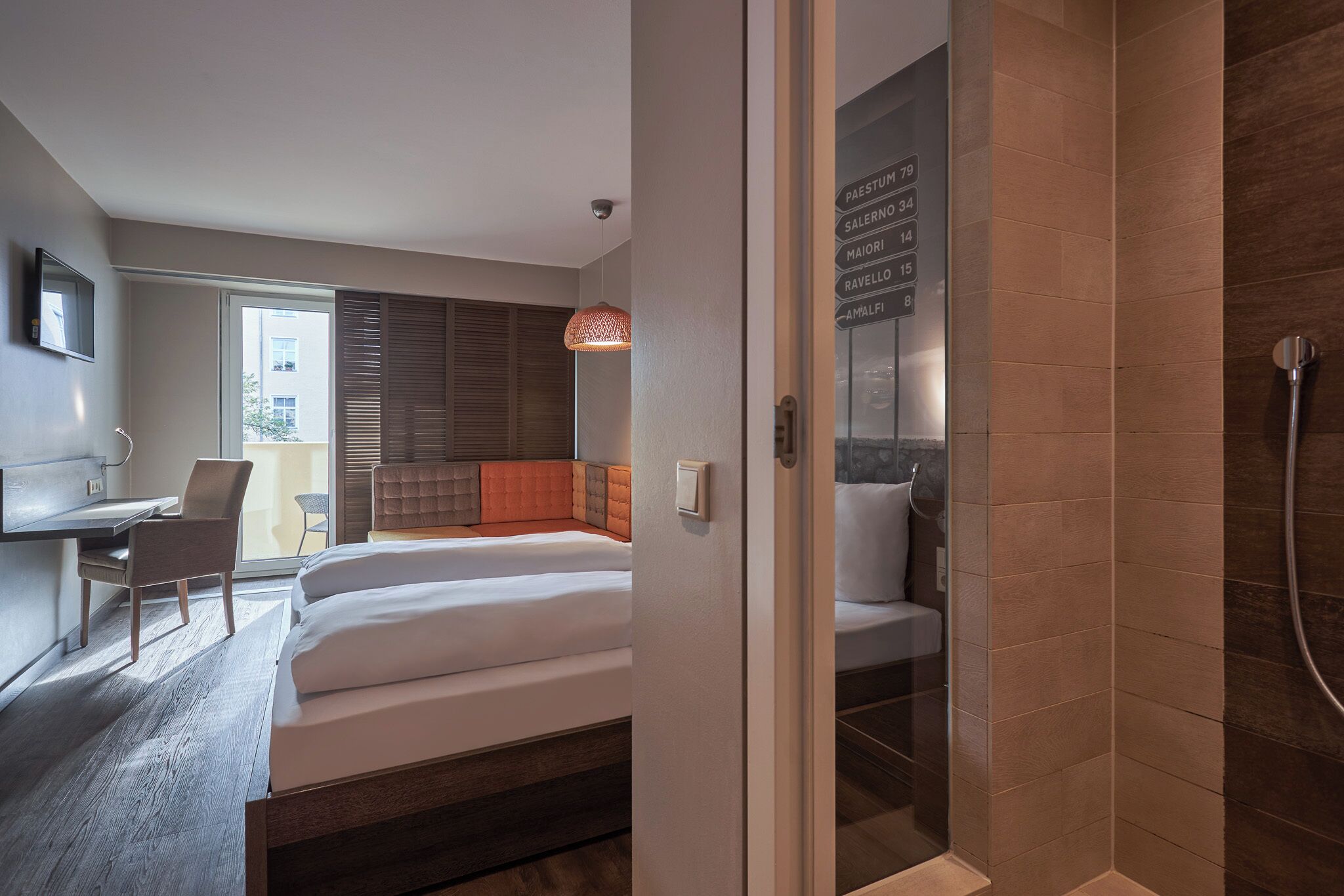 A modern hotel room with a bed, desk, chair, and a glass shower enclosure.