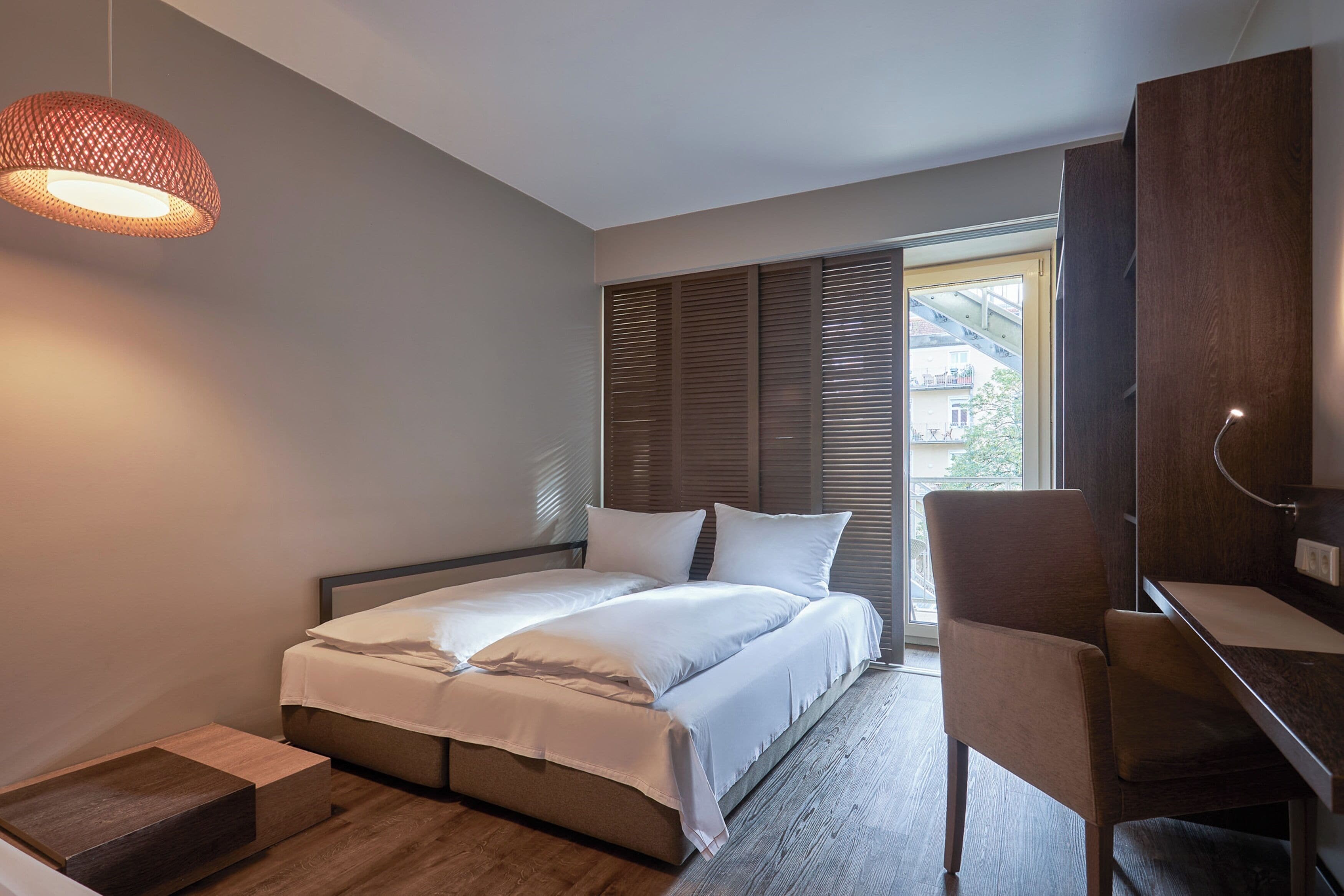 Un dormitorio moderno con cama, mesita de noche, silla y un amplio ventanal con persianas.