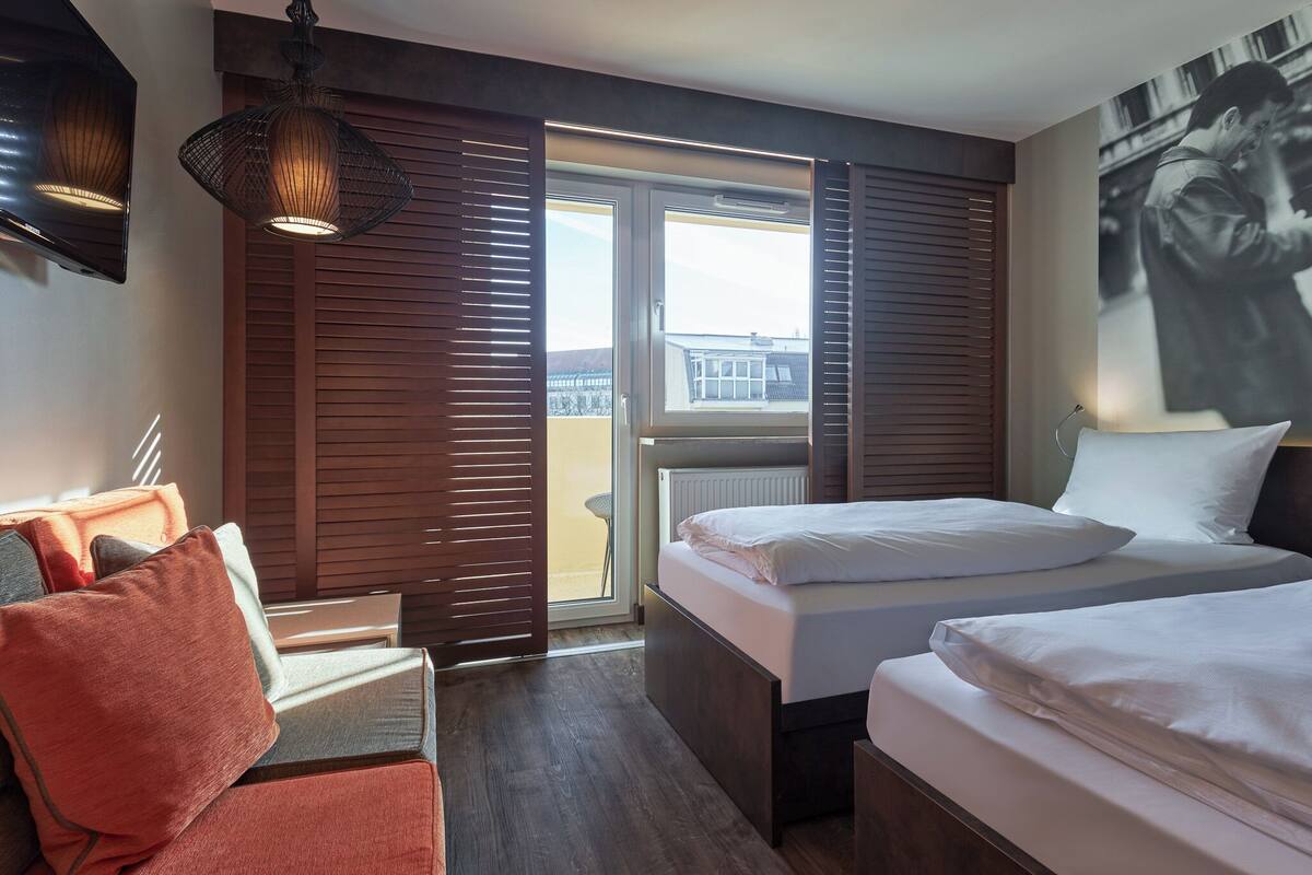 Habitación de hotel con dos camas, un sofá, un ventanal con persianas y una obra de arte colgada en la pared.