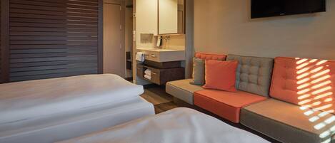 Habitación de hotel con dos camas, un sofá, una televisión y un baño con espejo y lavamanos.