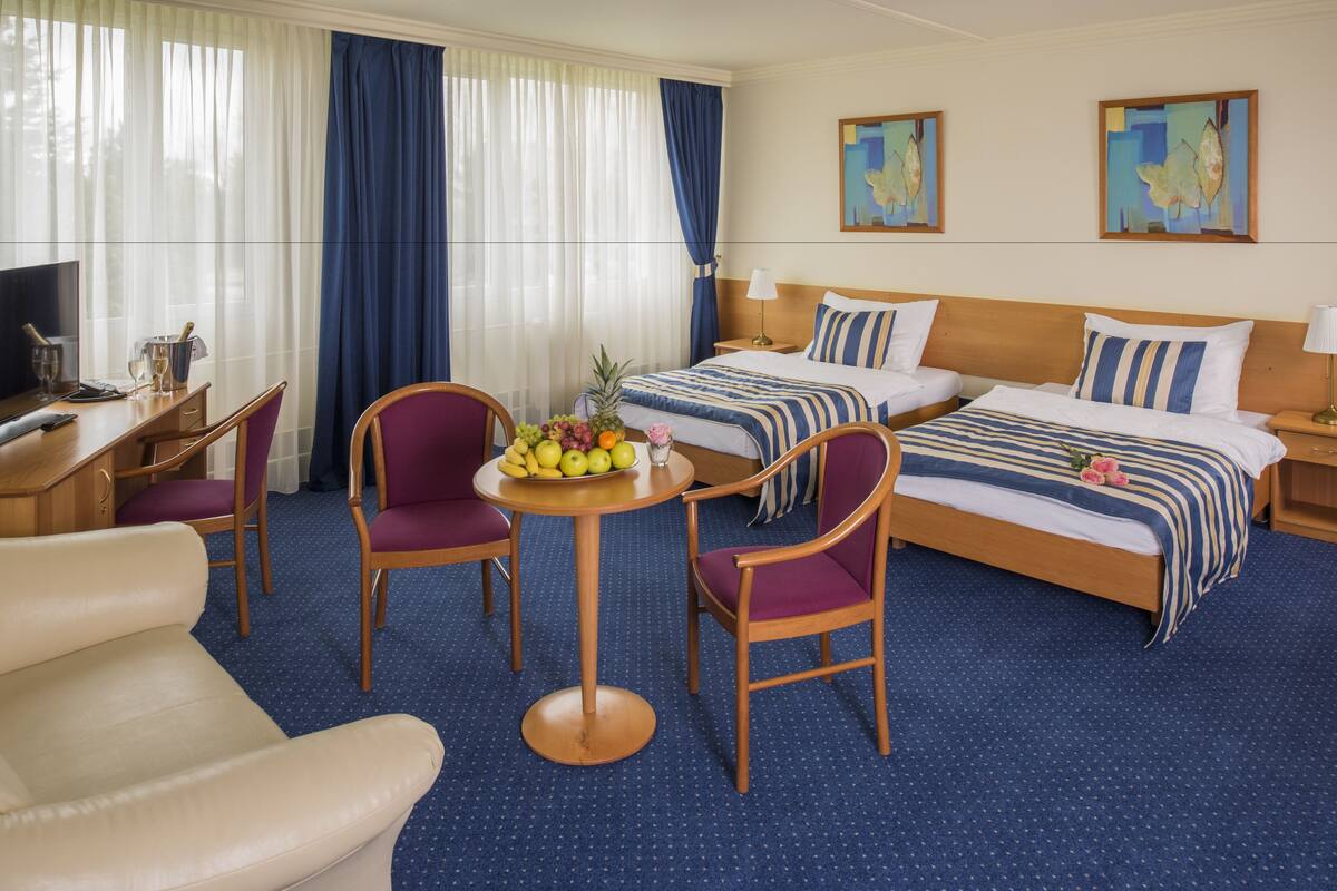 Habitación de hotel con dos camas, un escritorio con computadora, dos sillas, una mesita con fruta y obras de arte en las paredes.