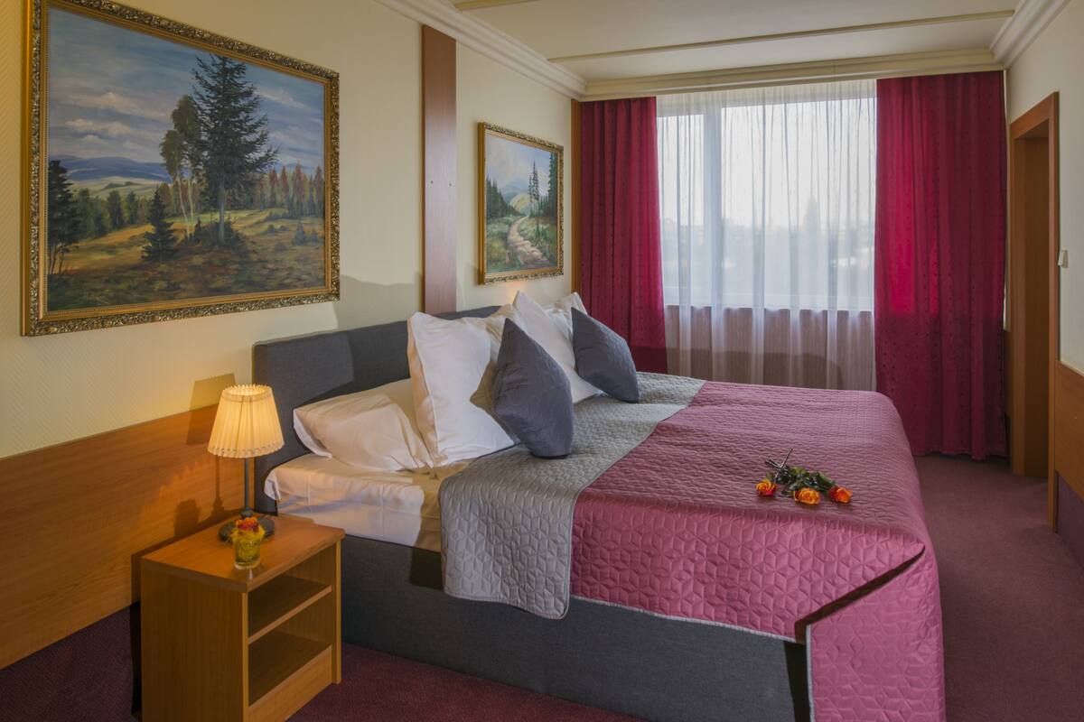 Habitación de hotel con cama, mesita de noche, lámpara, dos cuadros y una ventana con cortinas.