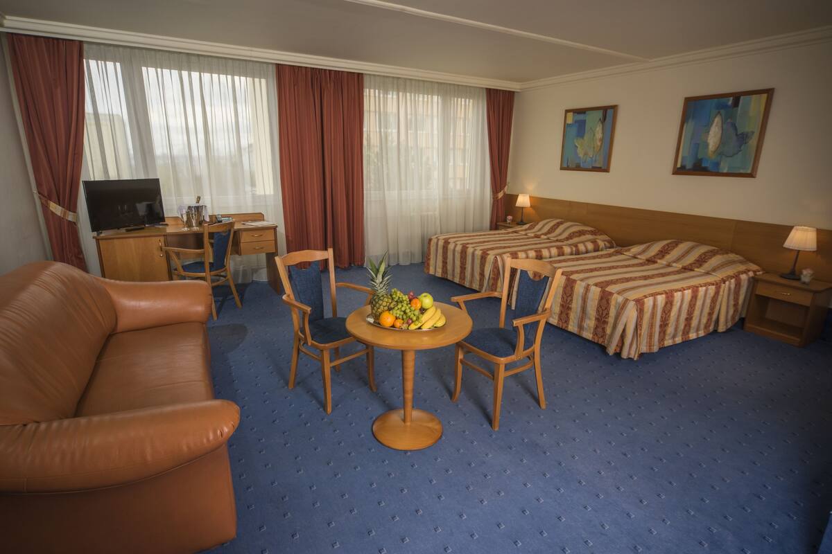 Habitación de hotel con sofá de cuero marrón, dos camas individuales, un escritorio con ordenador y un frutero sobre una mesita.