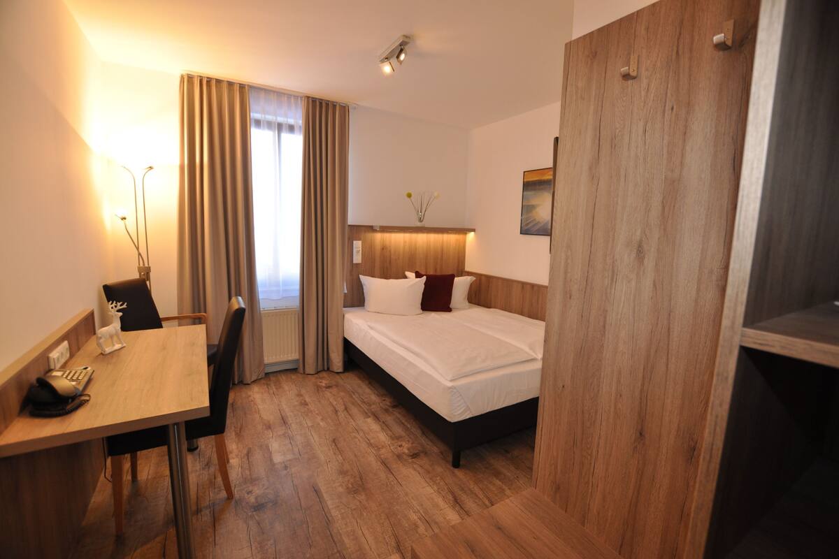 Habitación de hotel con piso de madera, cama con sábanas blancas, escritorio con teléfono y ventana con cortinas.
