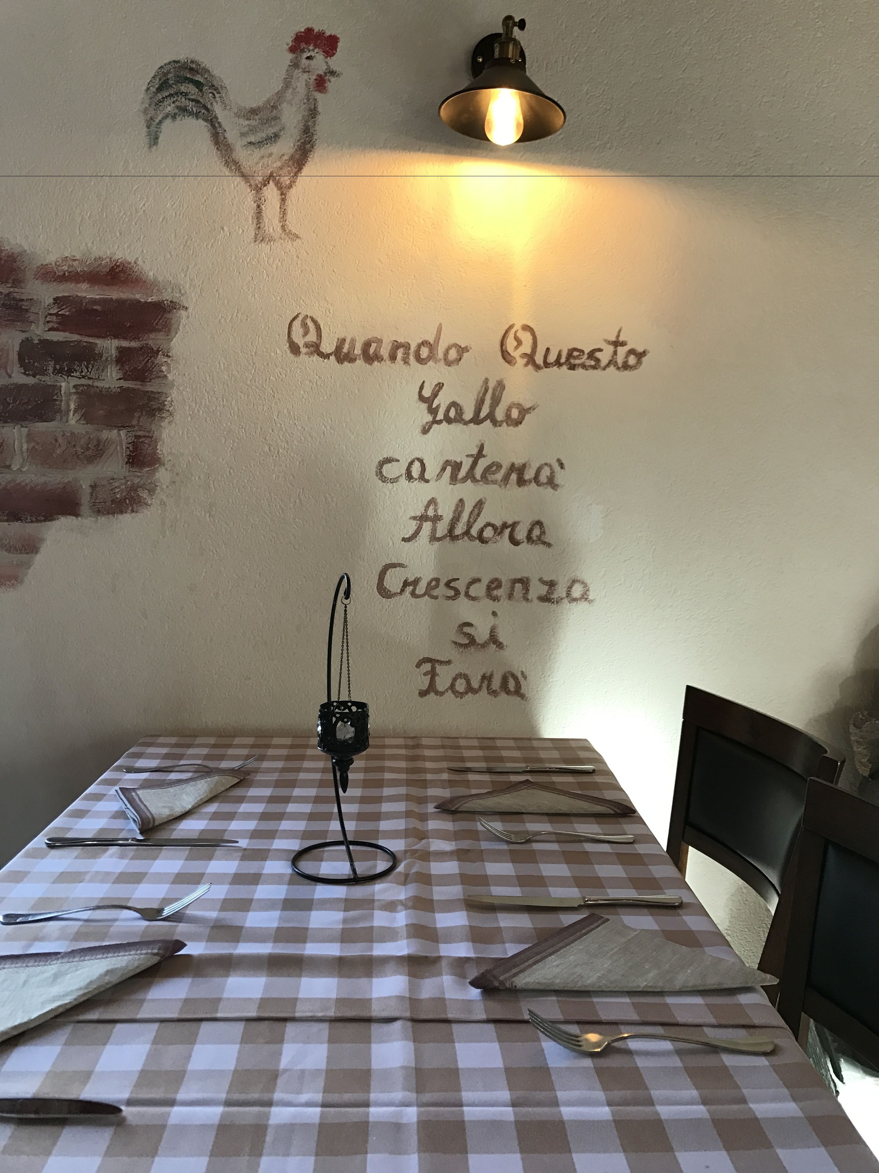 A dining area with a checkered tablecloth, a wall-mounted light fixture, and a wall decoration featuring a rooster and a phrase in Italian.