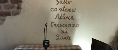 A dining area with a checkered tablecloth, a wall-mounted light fixture, and a wall decoration featuring a rooster and a phrase in Italian.
