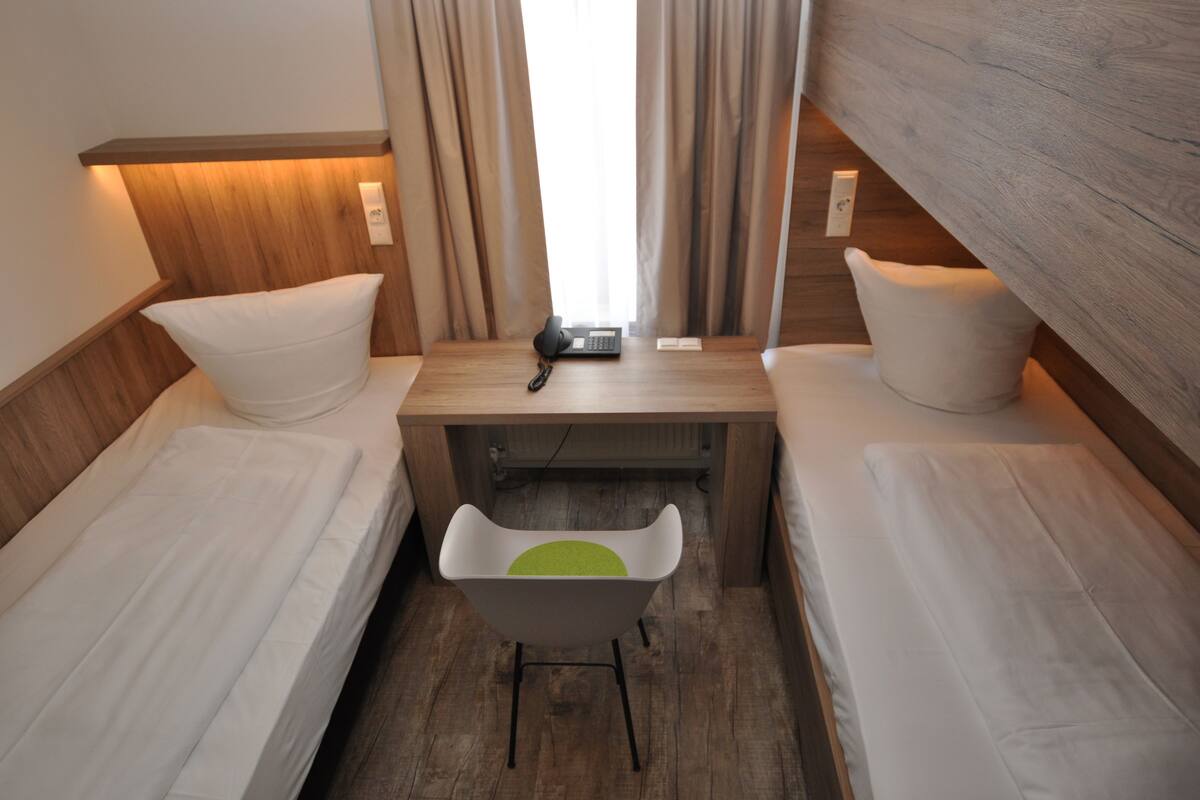 Habitación de hotel compacta con dos camas individuales, una mesita con teléfono y una silla.