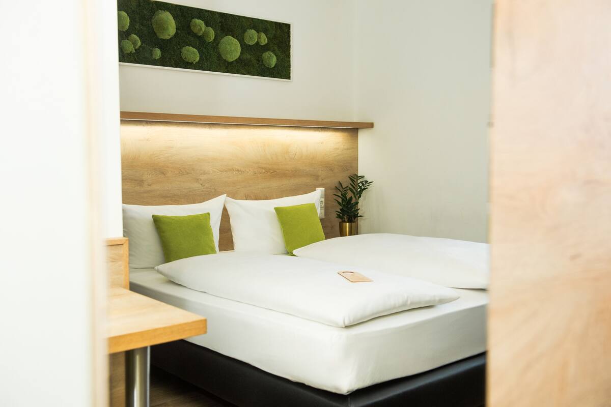 Una cama bien hecha con sábanas blancas y almohadas verdes, un cabecero de madera y una pequeña planta en maceta sobre la mesita de noche.
