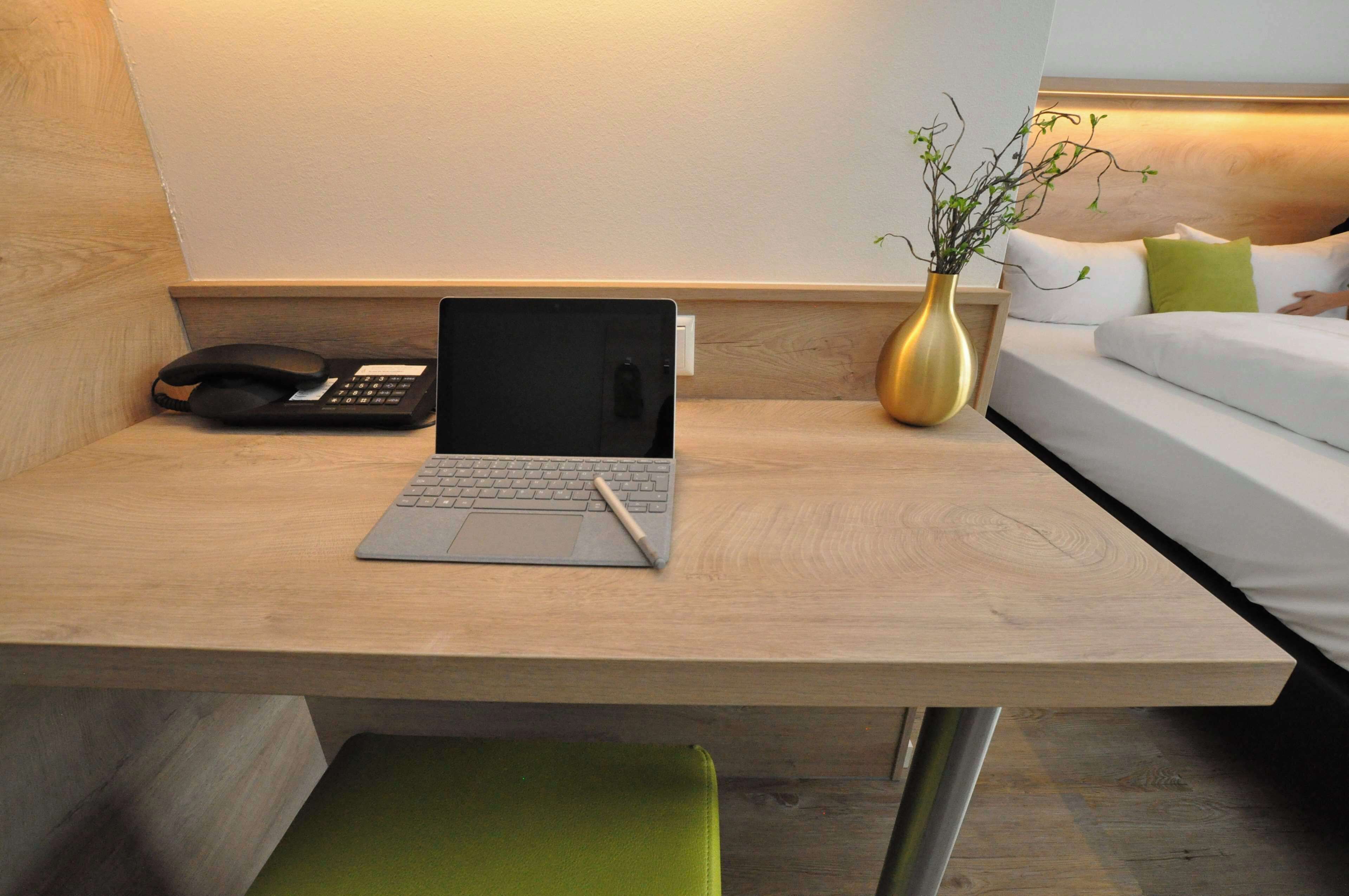 Habitación de hotel con escritorio de madera, una computadora portátil, un teléfono, un florero con planta y una cama con almohadas blancas y verdes.
