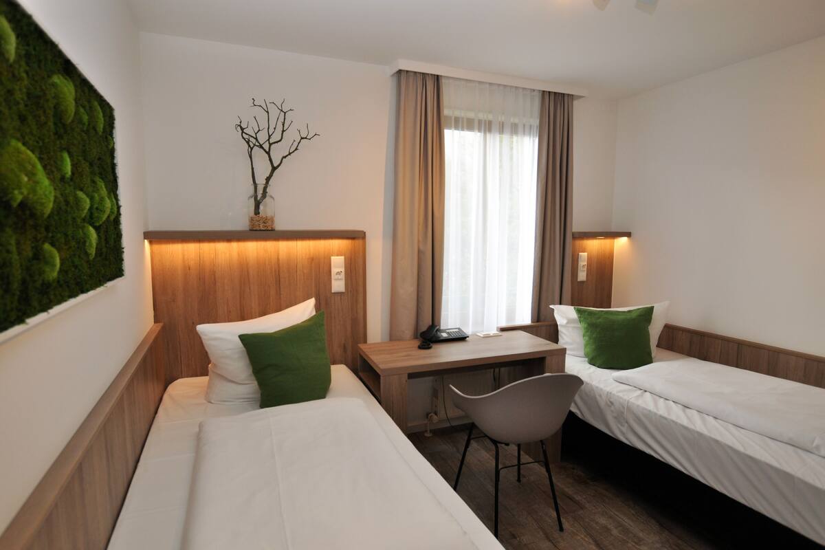 Habitación de hotel con dos camas individuales, un escritorio, una silla, una mesita pequeña y decoración en la pared.