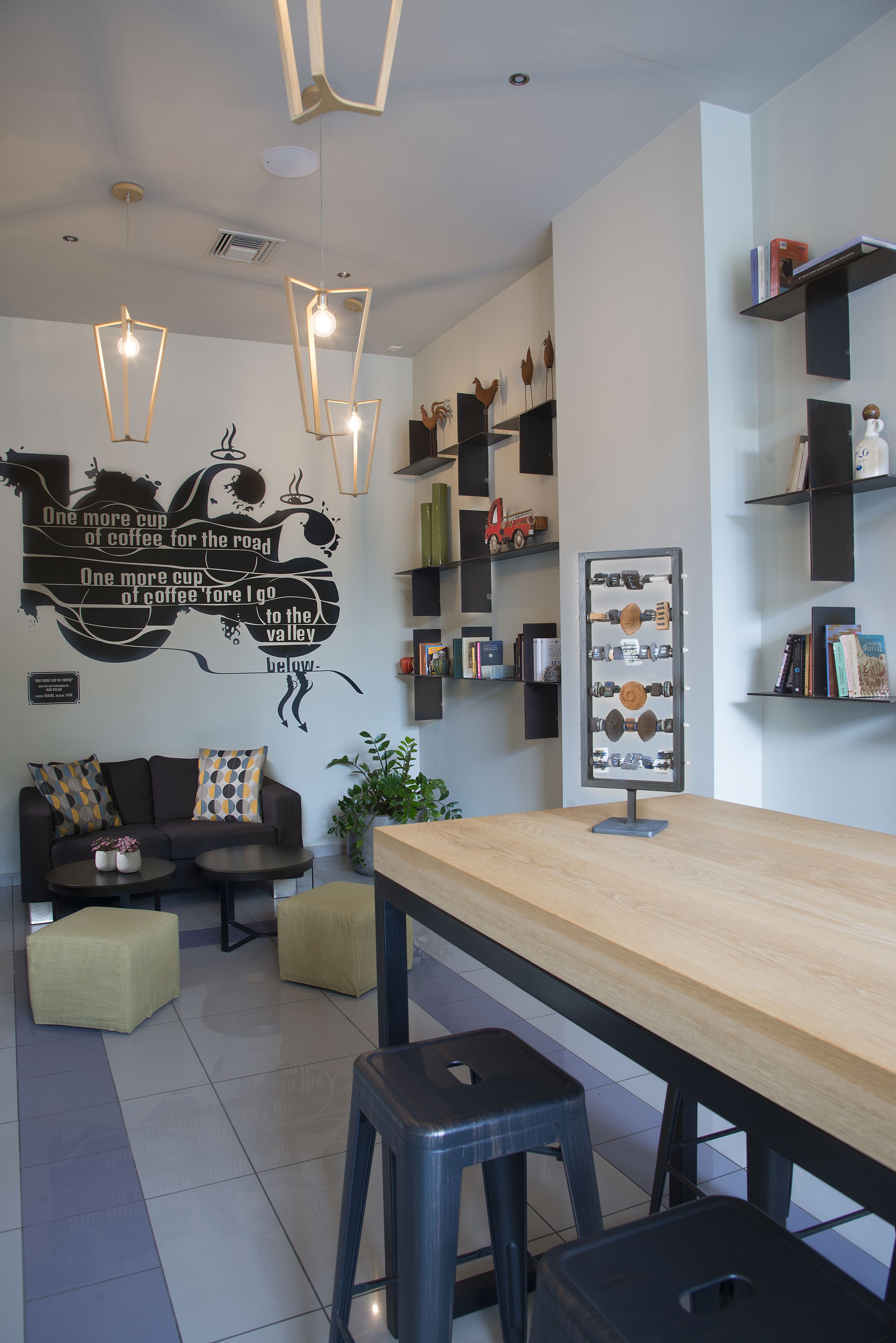 A modern cafe interior with a wooden table, black stools, a wall-mounted coffee display, and decorative shelves.