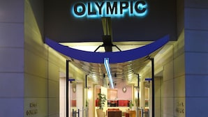La entrada del Hotel Olímpico, con un diseño moderno, presenta una puerta de vidrio, un área de recepción visible a través del vidrio y plantas en macetas en la entrada.