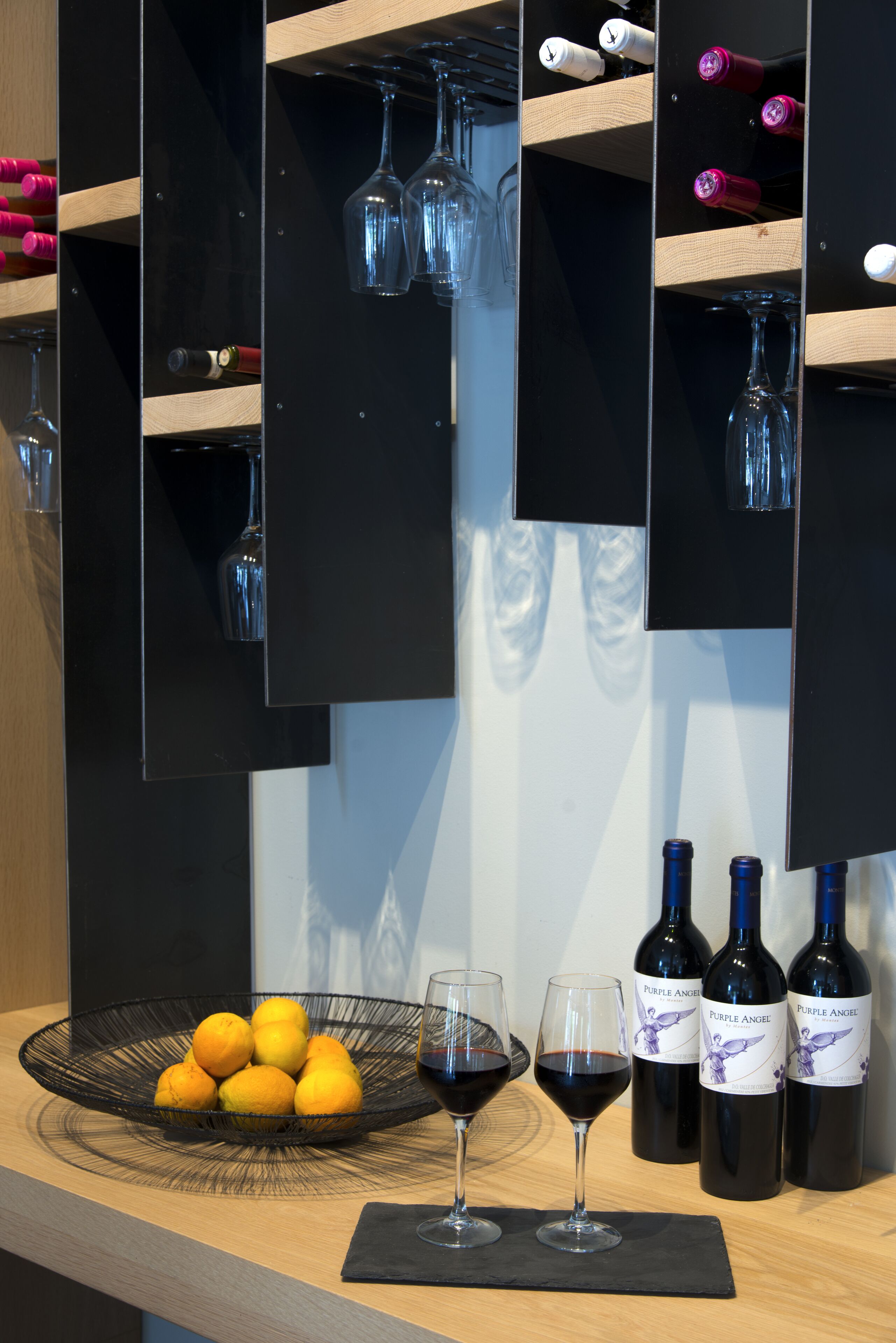 A wine rack with bottles and glasses, a bowl of lemons, and a wine glass on a wooden counter.